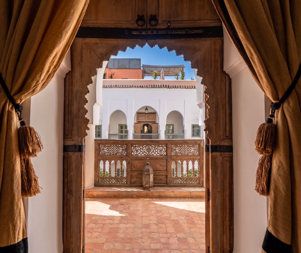 Vue depuis l'entrée d'un riad traditionnel marocain géré par Conciera — porte en cèdre sculpté, rideaux à pompons, arche moucharabieh donnant sur le patio intérieur, balcons en bois ajouré et sol en terre cuite